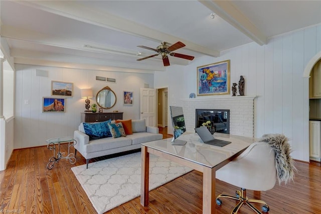living area with a ceiling fan, visible vents, a brick fireplace, wood finished floors, and beamed ceiling