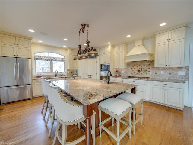 kitchen featuring premium range hood, a center island with sink, hanging light fixtures, appliances with stainless steel finishes, and light hardwood / wood-style floors