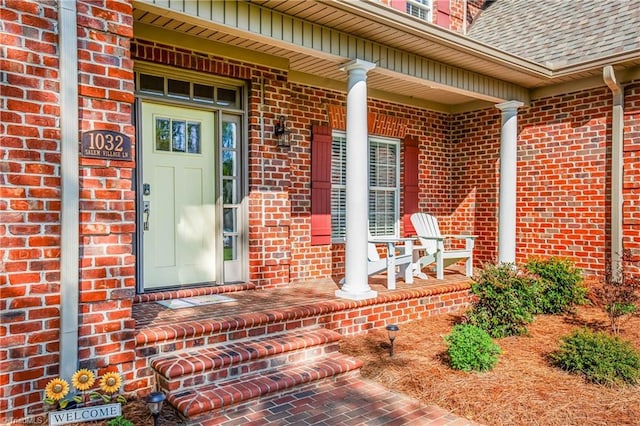 entrance to property featuring brick siding, a shingled roof, and covered porch
