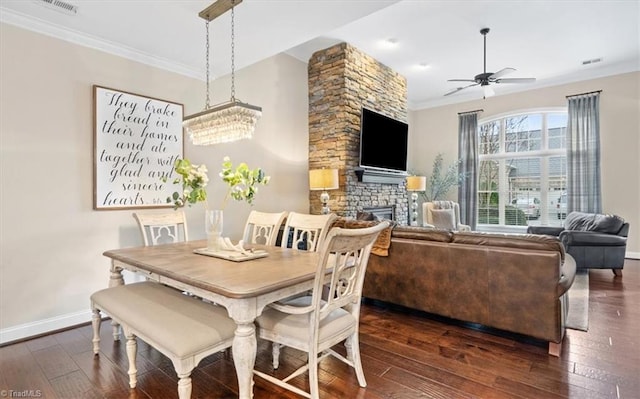 dining area featuring ceiling fan with notable chandelier, a stone fireplace, ornamental molding, and dark wood-type flooring