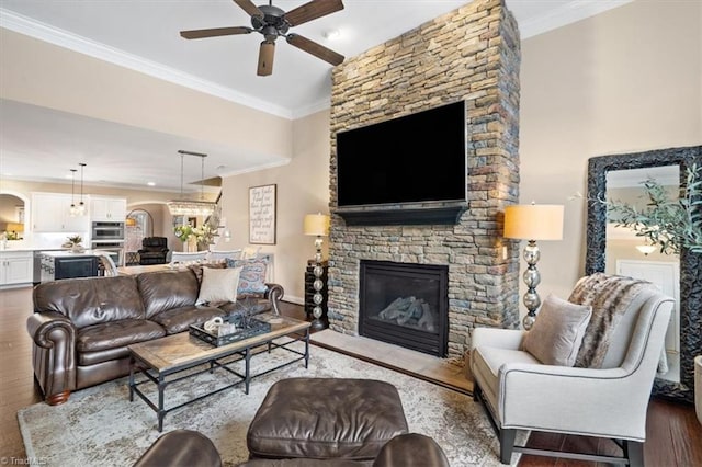 living room with ceiling fan with notable chandelier, light wood-type flooring, a stone fireplace, and crown molding