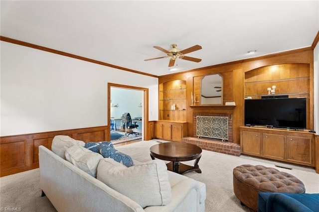 carpeted living area with crown molding, ceiling fan, built in features, a wainscoted wall, and a fireplace
