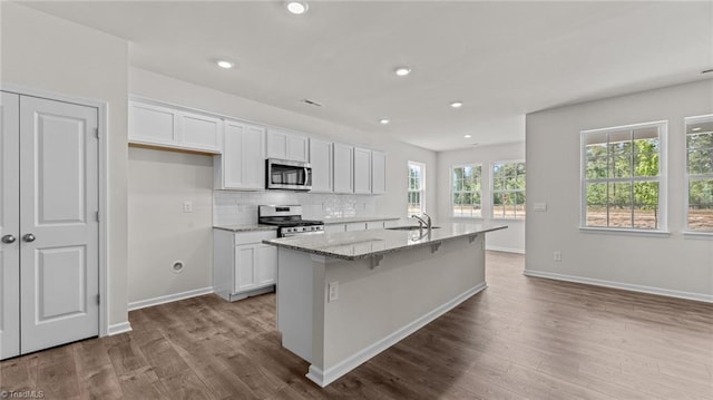 kitchen featuring appliances with stainless steel finishes, white cabinets, light wood-type flooring, light stone countertops, and an island with sink