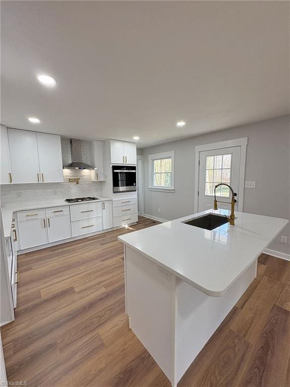 kitchen with oven, wood finished floors, gas stovetop, a sink, and wall chimney exhaust hood