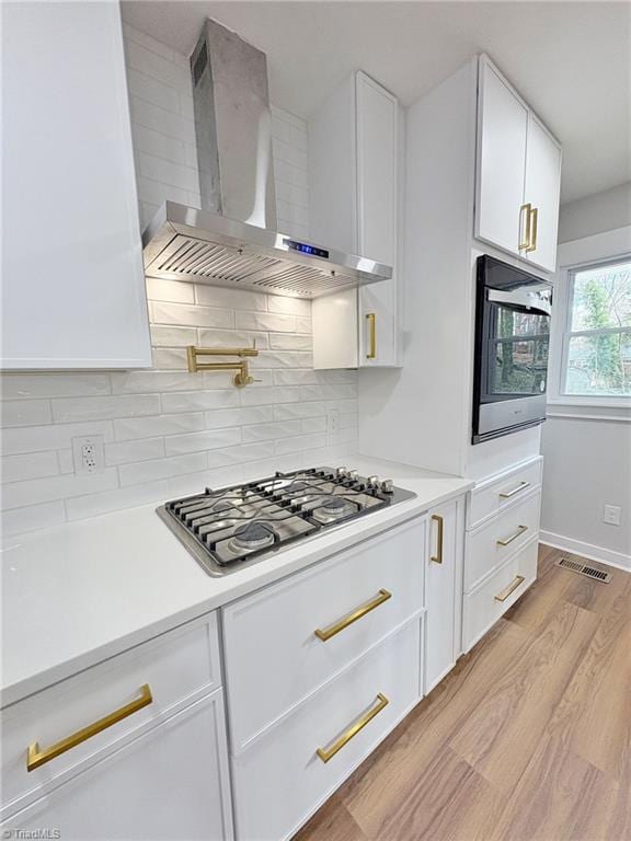 kitchen featuring visible vents, oven, wall chimney range hood, stainless steel gas stovetop, and white cabinetry