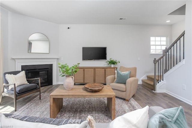 living room featuring a glass covered fireplace, baseboards, wood finished floors, visible vents, and stairway