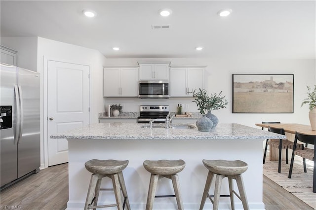 kitchen featuring light wood-type flooring, light stone counters, stainless steel appliances, white cabinets, and an island with sink