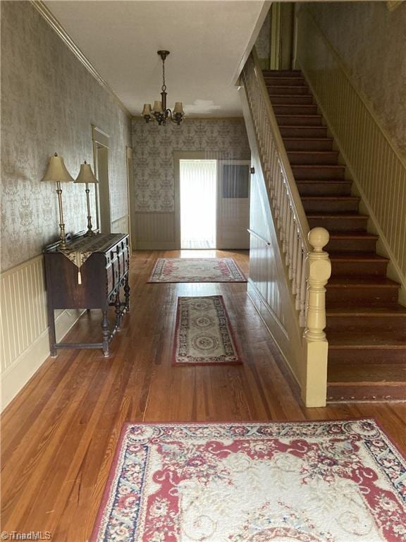 foyer entrance with a wainscoted wall, wood-type flooring, stairway, a chandelier, and wallpapered walls