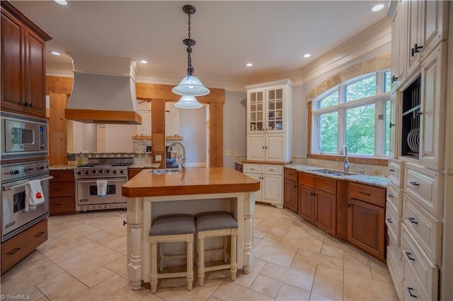 kitchen featuring a kitchen island with sink, custom range hood, decorative columns, butcher block countertops, and appliances with stainless steel finishes