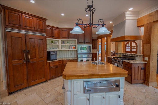 kitchen with decorative light fixtures, built in appliances, butcher block counters, sink, and premium range hood