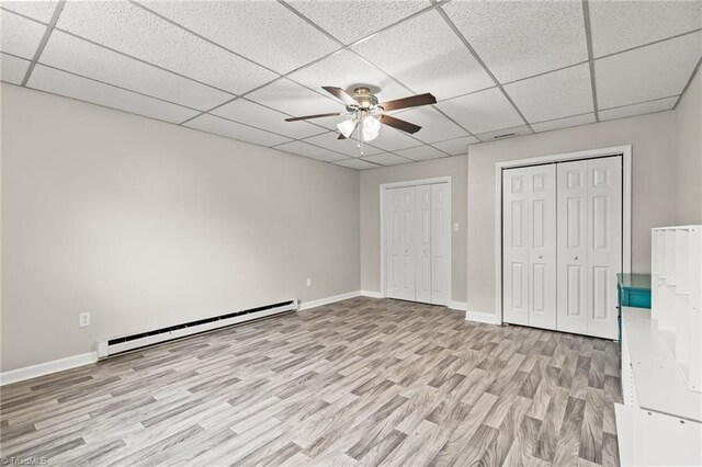 unfurnished bedroom featuring multiple closets, ceiling fan, a baseboard heating unit, light wood-type flooring, and a drop ceiling