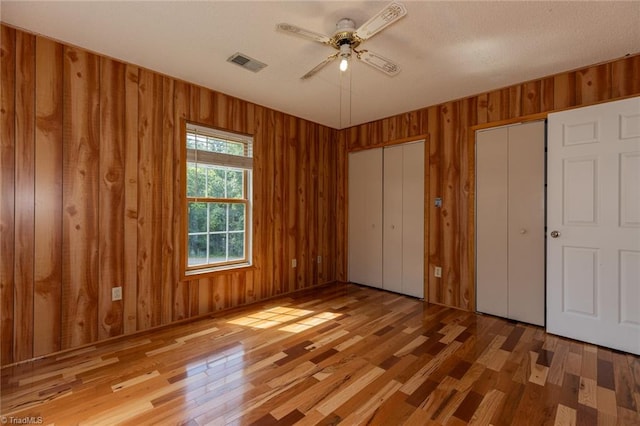 unfurnished bedroom with two closets, wooden walls, light wood-type flooring, and ceiling fan