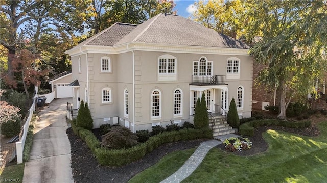 georgian-style home with a front yard, a shingled roof, a balcony, concrete driveway, and stucco siding