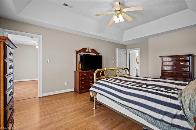 bedroom with light wood-type flooring, ensuite bathroom, a raised ceiling, and ceiling fan