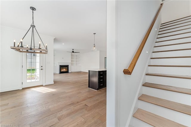 staircase with ceiling fan with notable chandelier, wood finished floors, a lit fireplace, and ornamental molding