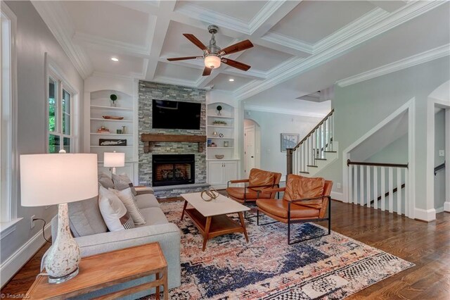 living room featuring ornamental molding, wood finished floors, a ceiling fan, a stone fireplace, and beam ceiling