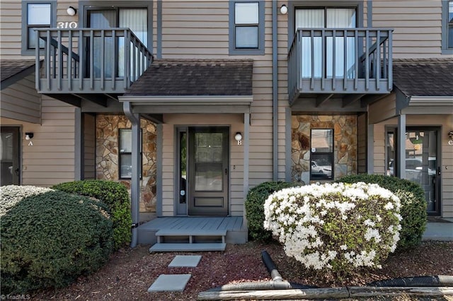 view of exterior entry featuring a shingled roof, stone siding, and a balcony