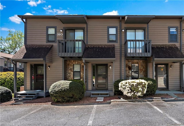 townhome / multi-family property featuring uncovered parking, roof with shingles, stone siding, and a balcony