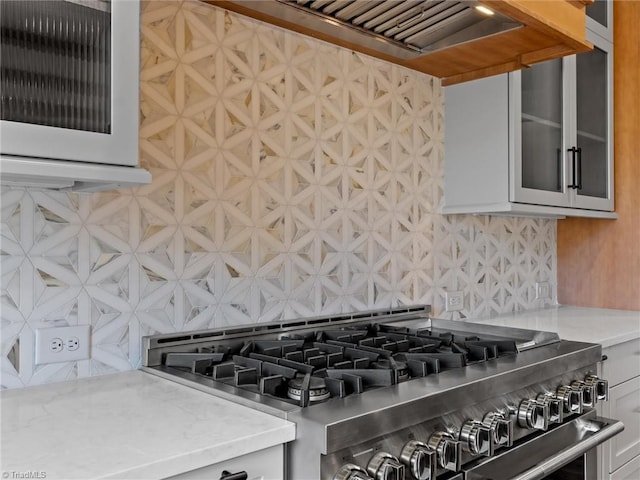 kitchen with tasteful backsplash, wall chimney exhaust hood, glass insert cabinets, light stone countertops, and high end stove