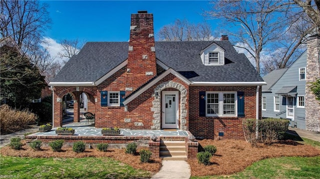 tudor house with brick siding, a patio area, a chimney, and roof with shingles