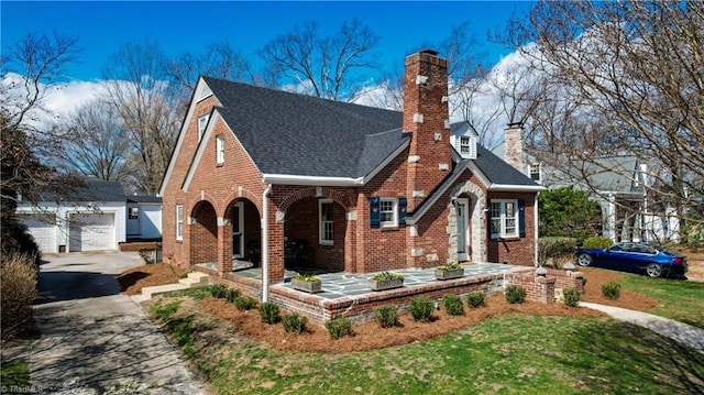 tudor-style house featuring a detached garage, roof with shingles, an outdoor structure, brick siding, and a chimney