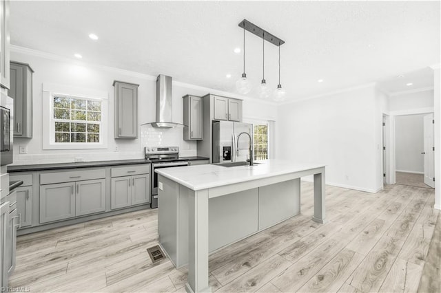 kitchen featuring decorative light fixtures, sink, wall chimney range hood, stainless steel appliances, and a center island with sink