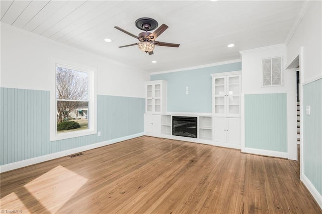 unfurnished living room with visible vents, wainscoting, a glass covered fireplace, ornamental molding, and wood finished floors