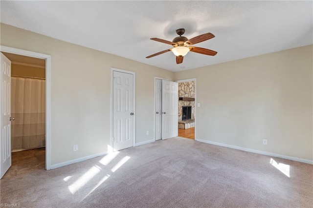 unfurnished bedroom with baseboards, ceiling fan, carpet, a stone fireplace, and two closets