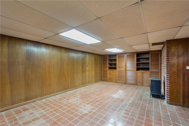 basement featuring built in features, a paneled ceiling, a wood stove, and wood walls