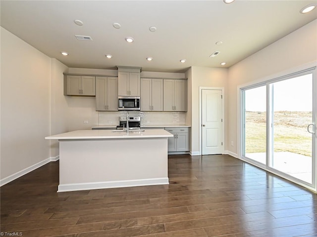 kitchen with gray cabinetry, stainless steel appliances, sink, dark hardwood / wood-style floors, and an island with sink