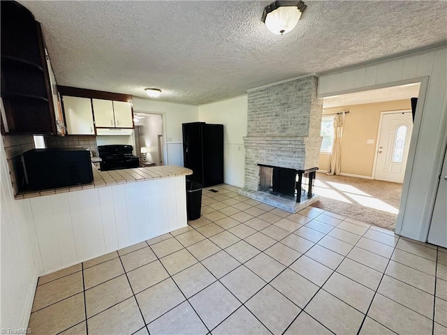kitchen with a brick fireplace, a peninsula, under cabinet range hood, tile counters, and black appliances