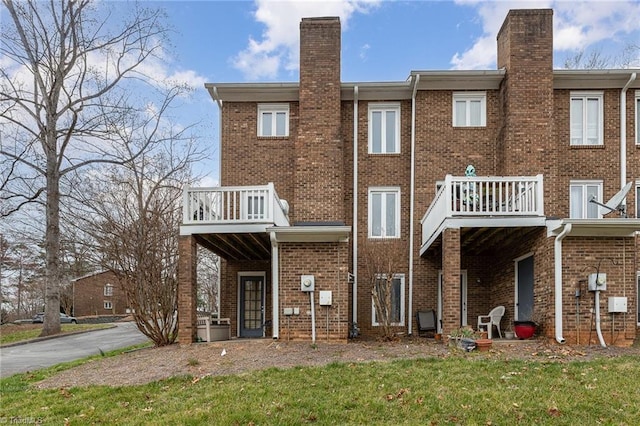 rear view of house with brick siding, a balcony, a chimney, and a lawn