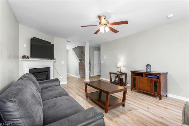 living area featuring a ceiling fan, light wood-type flooring, baseboards, a glass covered fireplace, and stairs