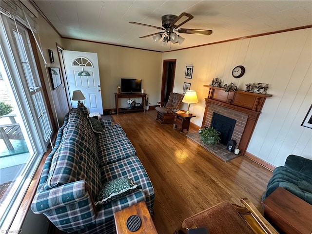 living area with a ceiling fan, wood finished floors, baseboards, ornamental molding, and a brick fireplace