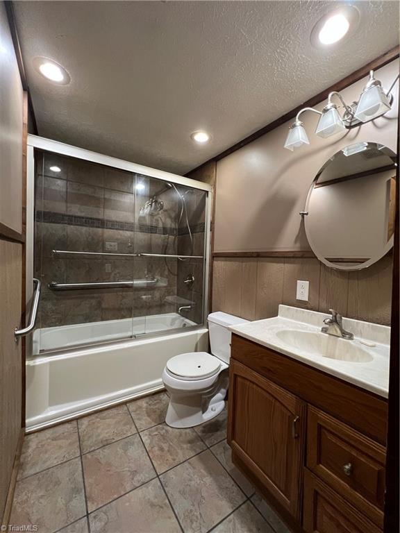 bathroom featuring a wainscoted wall, toilet, combined bath / shower with glass door, a textured ceiling, and vanity