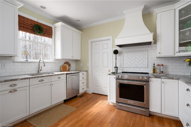 kitchen featuring premium range hood, ornamental molding, appliances with stainless steel finishes, white cabinets, and a sink