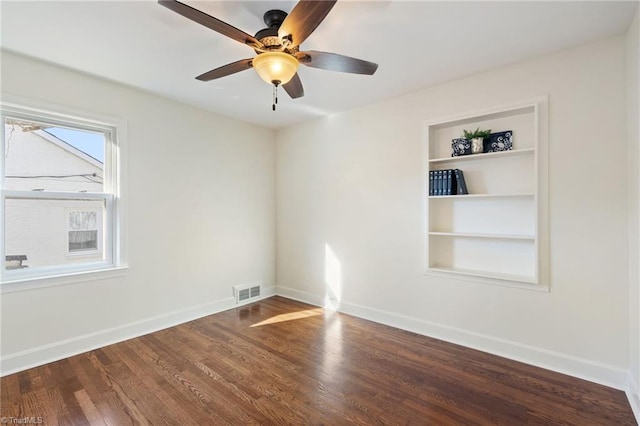 empty room with visible vents, built in shelves, wood finished floors, baseboards, and ceiling fan