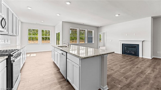 kitchen with appliances with stainless steel finishes, an island with sink, sink, light stone counters, and light hardwood / wood-style flooring