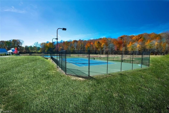 view of tennis court featuring a forest view, a lawn, and fence