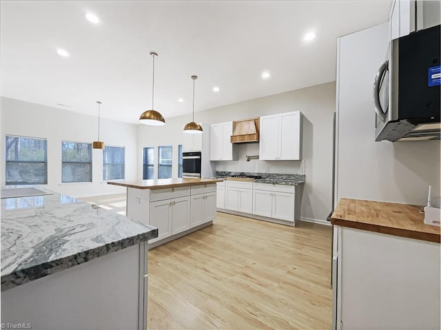 kitchen with premium range hood, butcher block countertops, white cabinetry, light wood finished floors, and stainless steel microwave