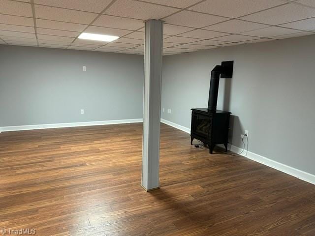 basement featuring dark wood-type flooring, a drop ceiling, and a wood stove