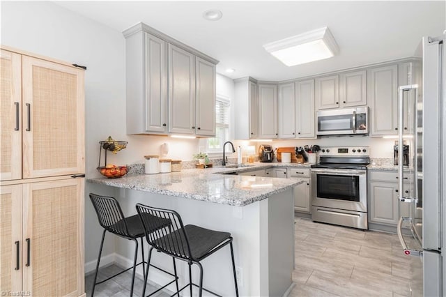kitchen featuring appliances with stainless steel finishes, a sink, a peninsula, a kitchen bar, and gray cabinetry