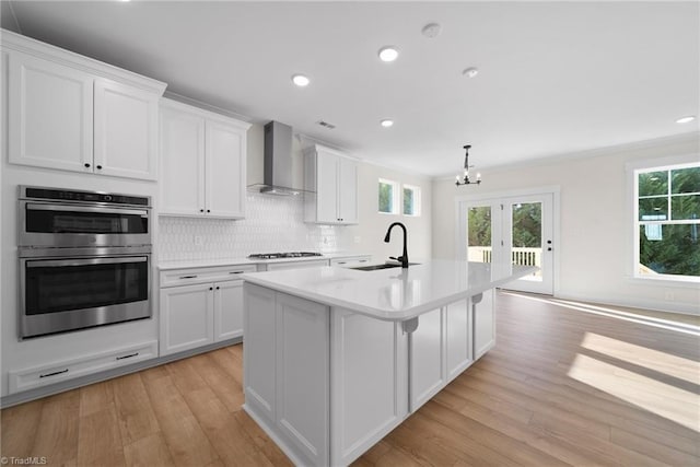 kitchen with wall chimney range hood, a sink, stainless steel double oven, gas stovetop, and light wood-type flooring