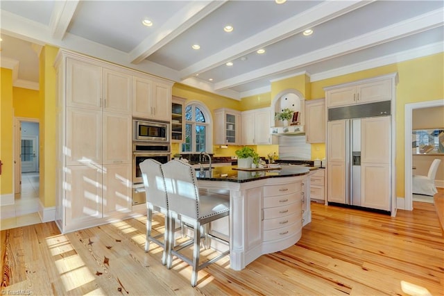 kitchen with a breakfast bar area, built in appliances, dark stone countertops, beam ceiling, and a kitchen island with sink