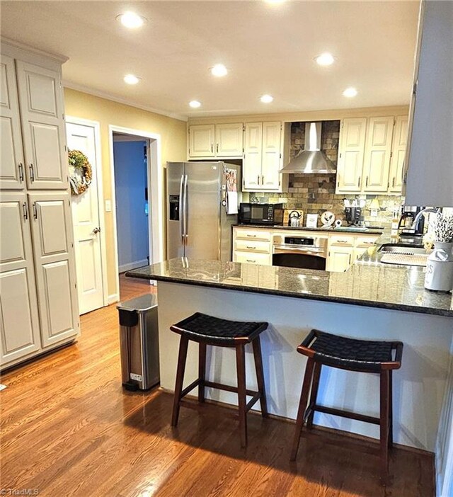 kitchen with appliances with stainless steel finishes, wood finished floors, a peninsula, wall chimney range hood, and backsplash