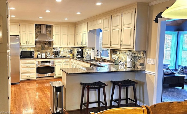 kitchen featuring stainless steel appliances, a sink, light wood-type flooring, a peninsula, and wall chimney exhaust hood