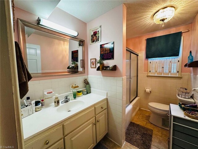 full bathroom with a textured ceiling, combined bath / shower with glass door, vanity, toilet, and tile walls