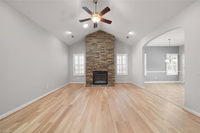 unfurnished living room with light wood finished floors, arched walkways, a stone fireplace, ceiling fan with notable chandelier, and baseboards