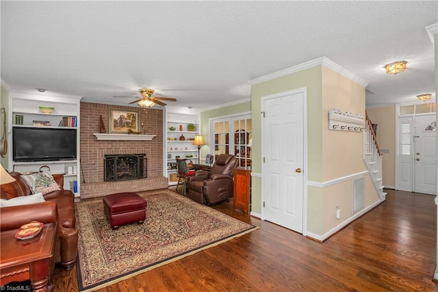 living room with built in shelves, ceiling fan, dark wood-type flooring, a fireplace, and ornamental molding