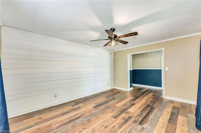 spare room featuring wood finished floors, wood walls, ceiling fan, baseboards, and crown molding